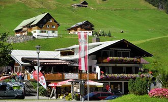 A charming inn with blooming balconies in a green mountain landscape. In the background, more traditional cottages can be seen. | © Cafe Baad Grund | Roman Schuster