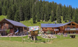 A picturesque mountain village with traditional wooden houses and a green meadow. Surrounded by tall trees and clear skies. | © Naturzeithof Café für dich | Stefanie Conticello