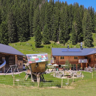 A picturesque mountain village with traditional wooden houses and a green meadow. Surrounded by tall trees and clear skies. | © Naturzeithof Café für dich | Stefanie Conticello