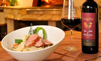 A delicious plate of meat and vegetables, served in a white bowl. Next to the dish, there is a glass of red wine and a bottle of wine on the table. | © Ristorante Trentino | Michael Monschau