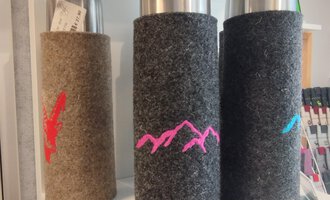 © Chrömle Schönes aus Filz | N. Lughammer Three thermos bottles with felt covers in different colors are standing on a shelf. The bottles have colored mountain motifs on the covers. | © Chrömle Schönes aus Filz | N. Lughammer