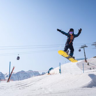 © OBERSTDORF · KLEINWALSERTAL BERGBAHNEN A snowboarder jumps over a snow hill in front of a clear mountain landscape. In the background, ski lifts and other skiers can be seen. | © OBERSTDORF · KLEINWALSERTAL BERGBAHNEN
