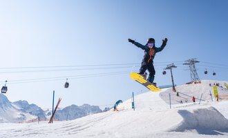 © OBERSTDORF · KLEINWALSERTAL BERGBAHNEN A snowboarder jumps over a snow hill in front of a clear mountain landscape. In the background, ski lifts and other skiers can be seen. | © OBERSTDORF · KLEINWALSERTAL BERGBAHNEN