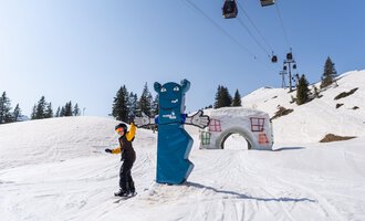 © OBERSTDORF · KLEINWALSERTAL BERGBAHNEN A skier stands on a snow-white slope next to a blue bear and an architectural element. In the background, lifts and snow-covered trees can be seen. | © OBERSTDORF · KLEINWALSERTAL BERGBAHNEN