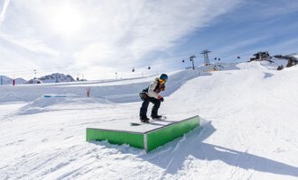 © OBERSTDORF · KLEINWALSERTAL BERGBAHNEN A snowboarder jumps over a green box in the snow. In the background, mountains and ski lifts are visible. | © OBERSTDORF · KLEINWALSERTAL BERGBAHNEN
