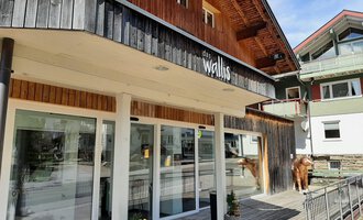 A modern building with wooden cladding stands in the foreground. There are large windows and a relaxed atmosphere in the surroundings. | © das Wallis Restaurant & Brasserie | Veronika Senn
