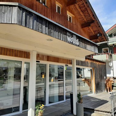 Ein modernes Gebäude mit Holzverkleidung steht im Vordergrund. Es gibt große Fenster und eine entspannte Atmosphäre in der Umgebung. | © das Wallis Restaurant & Brasserie | Veronika Senn