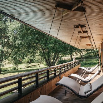 © Naturhotel Chesa Valisa | Heldentheater Eine entspannende Veranda mit modernen Liegen und Blick auf einen grünen Garten. Die Holzdecke und die natürliche Umgebung schaffen eine ruhige Atmosphäre. | © Naturhotel Chesa Valisa | Heldentheater