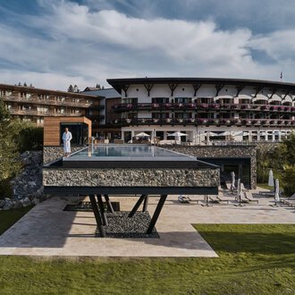 © Travel Charme Ifen Hotel Ein modernes Hotel mit einem großen, gläsernen Pool steht im Vordergrund. Die umliegende Landschaft ist grün und einladend. | © Travel Charme Ifen Hotel