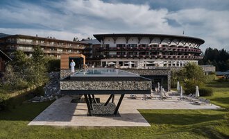 © Travel Charme Ifen Hotel Ein modernes Hotel mit einem großen, gläsernen Pool steht im Vordergrund. Die umliegende Landschaft ist grün und einladend. | © Travel Charme Ifen Hotel