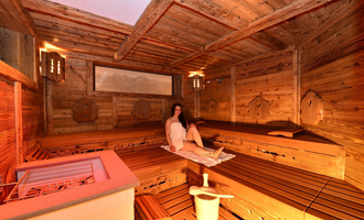 Eine gemütliche Sauna mit Holzwänden und Bänken. Eine entspannte Person sitzt im Raum und genießt die Wärme.