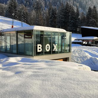 © Sport Kessler GmbH A modern glass building with the inscription "BOX" stands amidst a snow-covered landscape. In the background, snow-covered trees and additional buildings can be seen. | © Sport Kessler GmbH