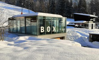 © Sport Kessler GmbH A modern glass building with the inscription "BOX" stands amidst a snow-covered landscape. In the background, snow-covered trees and additional buildings can be seen. | © Sport Kessler GmbH