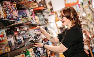 Eine Frau schaut sich ein LEGO-Set in einem Spielzeuggeschäft an. Sie lächelt und hält die Verpackung in der Hand.