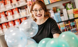 Eine junge Frau mit Brille lächelt und hält bunte Luftballons in einem Geschäft. Im Hintergrund sind Bücherregale und andere Artikel zu sehen.
