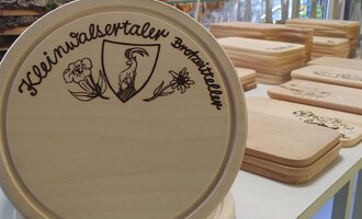 © Der Bunte Laden | Romana Volderauer Eine Holzplatte mit dem Aufdruck "Kleinwalsertaler Brotmitteller" ist im Vordergrund zu sehen. Im Hintergrund liegen weitere Holzplatten auf einem Tisch. | © Der Bunte Laden | Romana Volderauer