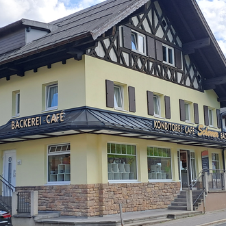 Ein hübsches Café mit einer Bäckerei in einem traditionellen Gebäude. Im Hintergrund sind Berge und ein blauer Himmel sichtbar. | © Kleinwalsertal Tourismus | Steffen Stöhr