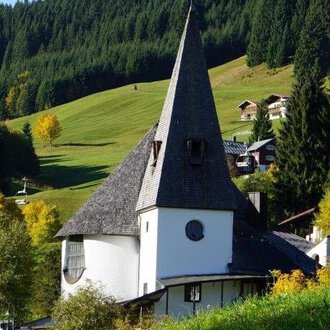 Eine malerische Kirche mit einem hohen spitzen Turm steht in einer grünen Landschaft. Im Hintergrund sind sanfte Hügel und einige Häuser zu sehen. | © Ev. Kirche Kleinwalsertal