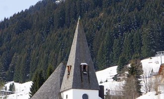 Eine charmante Kirche mit einem spitzen Turm in einer schneebedeckten Landschaft. Im Hintergrund sind grüne Berge zu sehen. | © Ev. Kirche Kleinwalsertal