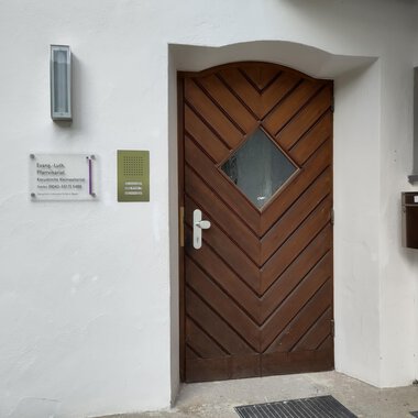 Eine braune Haustür mit einem rautenförmigen Fenster. Neben der Tür befindet sich ein Briefkasten und ein Schild mit Informationen. | © Ev. Kirche Kleinwalsertal