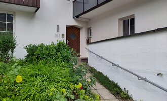 Ein Eingangsbereich eines Hauses mit einer grünen, blühenden Gartenlandschaft. Die Eingangstür ist aus Holz und hat eine einzigartige Form. | © Ev. Kirche Kleinwalsertal