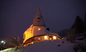 Ein gemütliches, schneebedecktes Haus mit einem spitzen Dach. Die Lichter im Gebäude strahlen eine warme Atmosphäre in der winterlichen Nacht aus. | © Ev. Kirche Kleinwalsertal
