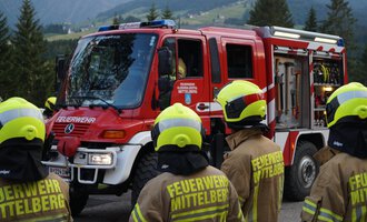 A fire truck is standing on a road, surrounded by firefighters in uniform yellow helmets. The landscape in the background shows trees and mountains. | © Feuerwehr Mittelberg | Christoph Hilbrand