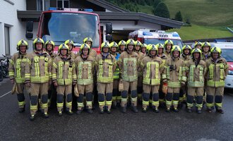 A group of firefighters in uniforms stands in front of a fire truck. In the background, a building and a green hill landscape can be seen. | © Feuerwehr Mittelberg | Christoph Hilbrand