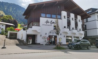 A modern building with a wooden and plaster facade that offers apartments. In the background, green mountains and a blue sky can be seen. | © Kleinwalsertal Tourismus | N. Lughammer