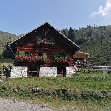 A rustic wooden house surrounded by green meadows and mountains. Flower boxes with colorful flowers adorn the windows. | © Fluchtalpe | N. Lughammer