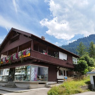 © Kleinwalsertal Tourismus | N. Lughammer Ein charmantes Haus mit einem blühenden Balkon in den Bergen. Die Umgebung ist grün und die Wolkenhimmel sorgt für eine schöne Atmosphäre. | © Kleinwalsertal Tourismus | N. Lughammer