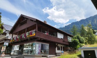 © Kleinwalsertal Tourismus | N. Lughammer Ein charmantes Haus mit einem blühenden Balkon in den Bergen. Die Umgebung ist grün und die Wolkenhimmel sorgt für eine schöne Atmosphäre. | © Kleinwalsertal Tourismus | N. Lughammer