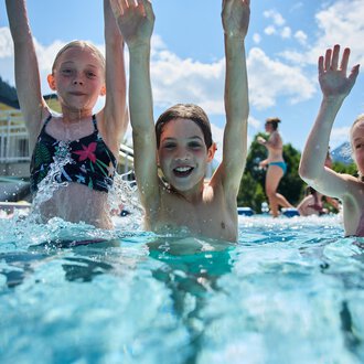 Three cheerful children are splashing in the swimming pool and having a lot of fun. They lift their hands in the air and enjoy the sun. | © Kleinwalsertal Tourismus | Oliver Farys