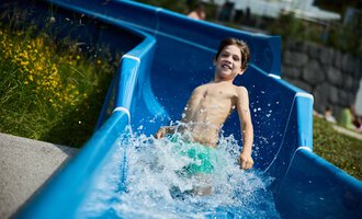 A boy happily slides down the water slide, splashing water in the process. In the background, there are green meadows and a few swimmers visible. | © Kleinwalsertal Tourismus | Oliver Farys