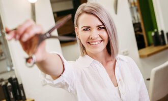 Eine fröhliche Friseurin hält eine Schere in der Hand und lächelt in die Kamera. Im Hintergrund sind Friseurelemente zu sehen. | © Friseur Haarwerkstatt | Franziska Diener