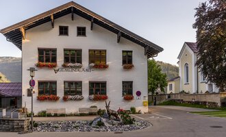 A charming community building with blooming flower boxes. In the background, additional buildings and trees can be seen. | © Kleinwalsertal Tourismus |  Steffen Berschin