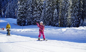 Ein kleines Mädchen in pinker Skiausrüstung fährt fröhlich auf Skiern und winkt. Im Hintergrund sieht man schneebedeckte Bäume und eine verschneite Piste. | © OBERSTDORF · KLEINWALSERTAL BERGBAHNEN