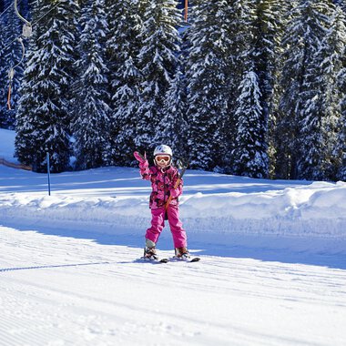 A little girl in pink ski gear is happily skiing and waving. In the background, you can see snow-covered trees and a snowy slope. | © OBERSTDORF · KLEINWALSERTAL BERGBAHNEN