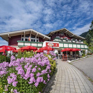 A cozy restaurant with a red facade and blooming flowers in the foreground. Red umbrellas provide shade for the guests. | © Gasthof Alpenblick | Theodor Pinn