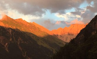 Eine beeindruckende Berglandschaft mit Sonnenuntergang, der die Felsen in warmen Orangetönen beleuchtet. Der Himmel ist bewölkt und sorgt für eine dramatische Atmosphäre. | © Gasthof Café Alpenwald | Thorsten Alpenwald