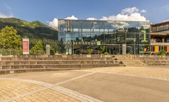 A modern building with a large glass facade, called Walserhaus. It is situated in a picturesque mountain landscape. | © Kleinwalsertal Tourismus | Steffen Berschin