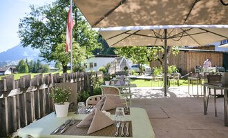 Eine einladende Terrasse mit gedeckten Tischen und Sonnenschirmen. Im Hintergrund sind grüne Bäume und eine schöne Landschaft zu sehen.