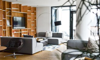 Ein modernes Wohnzimmer mit grauen Sofas und einem Holzregal. Große Fenster sorgen für viel natürliches Licht.