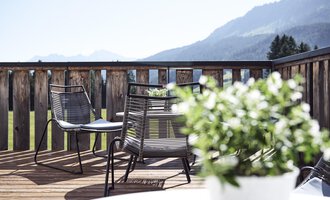 Eine gemütliche Terrasse mit zwei Stühlen und einem Tisch. Im Hintergrund sind Berge und ein klarer Himmel zu sehen.