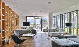 Ein modernes Wohnzimmer mit großen Fenstern und viel natürlichem Licht. Die Möbel sind stilvoll und minimalistisch, mit einem Bücherregal aus Holz und komfortablen Sitzbereichen.