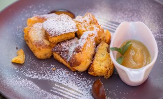 Ein Teller mit goldbraunen, zuckergestreuten Pfannkuchenstücken und einer Schüssel Apfelmus. Das Gericht sieht appetitlich aus und wird mit frischer Minze garniert.