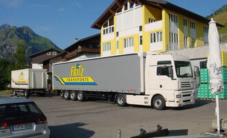 © Getränke Service Fritz | Helmut Fritz Ein Lkw von Fritz Transporte steht auf einem Parkplatz vor einem großen Gebäude. Im Hintergrund sind grüne Berge zu sehen. | © Getränke Service Fritz | Helmut Fritz