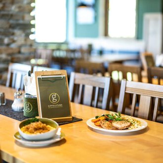 A cozy restaurant with wooden chairs and a clean table. On the table lies a menu, next to it is a plate with meat and vegetables as well as a bowl of rice. | © OBERSTDORF·KLEINWALSERTAL BERGBAHNEN