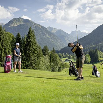 Eine Gruppe von Golfern übt auf einem malerischen Golfplatz mit Bergen im Hintergrund. Die Sonne scheint und die Landschaft ist grün und einladend. | © Golfclub Oberstdorf | ©kunst.joachimweiler.de