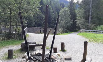 Eine Feuerstelle in einem Waldgebiet, umgeben von Bäumen und einem Kiesweg. Im Hintergrund sind Holzstämme und eine Brücke zu sehen. | © Kleinwalsertal Tourismus | Veronika Senn